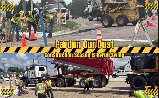 Two images show construction workers in yellow vests and hard hats performing road work. The top image shows workers shoveling dirt, and the bottom image shows workers spreading gravel from a large red dump truck. Yellow and black "Pardon Our Dust" and "Construction Season Is In Full Swing" text overlays are present.